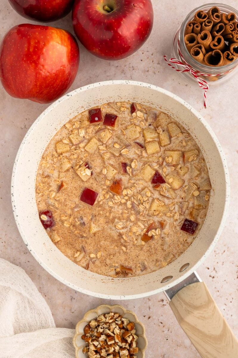 Overhead view of rolled oats cooking in a pot of liquid with apples, cinnamon, and chopped nuts.