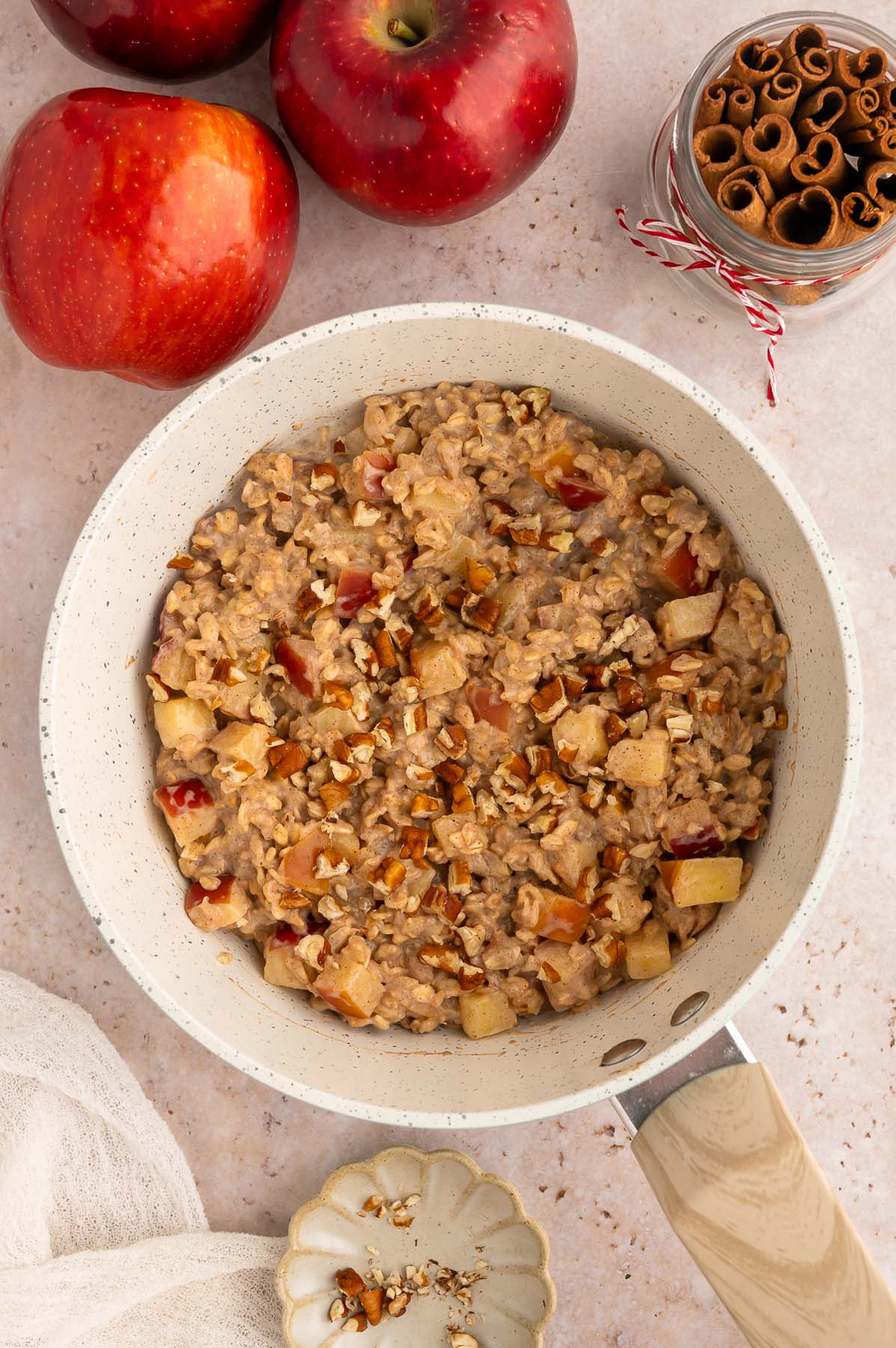 Apple oatmeal in a bowl, viewed from above. Two red apples and a jar of cinnamon sticks sit near the bowl of cereal.