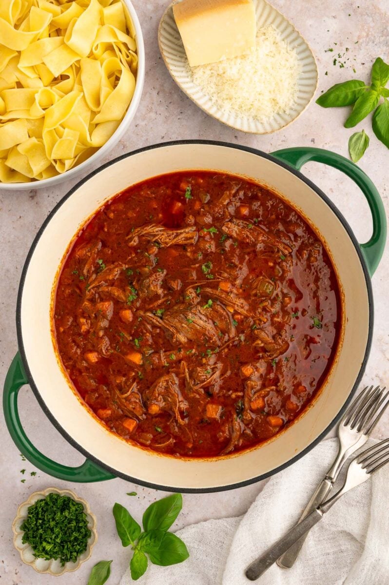 Fully cooked chuck roast pasta sauce in a dutch oven with a bowl of pasta noodles and parmesan on the side.