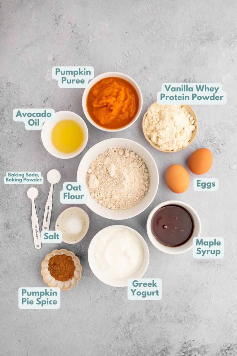 Ingredients for pumpkin protein muffins in small bowls: pumpkin puree, vanilla whey protein powder, eggs, maple syrup, greek yogurt, oat flour, avocado oil, baking powder, baking soda, salt, pumpkin pie spice.