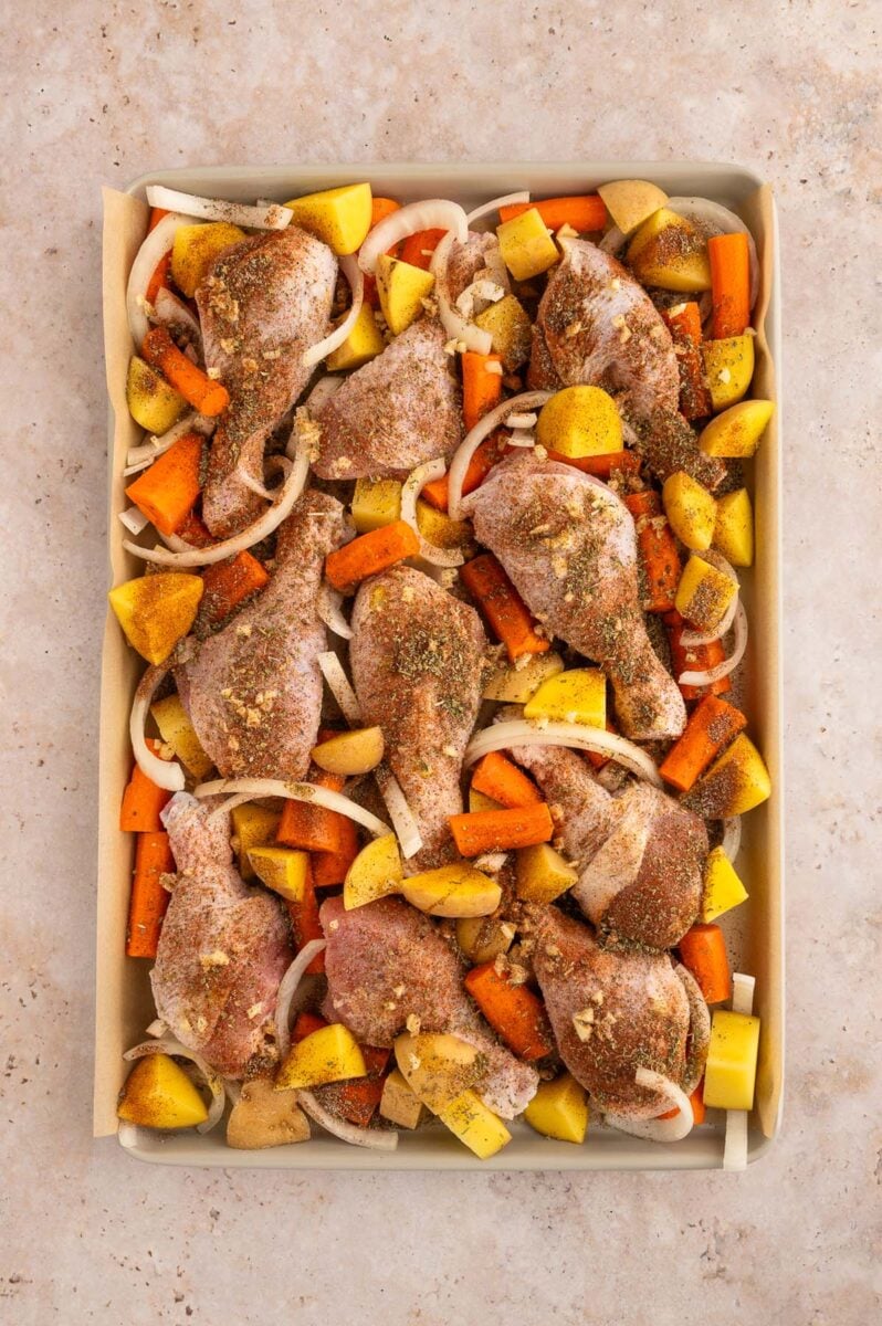 Uncooked chicken drumstick on top of potatoes, carrots and onions on a sheet pan.
