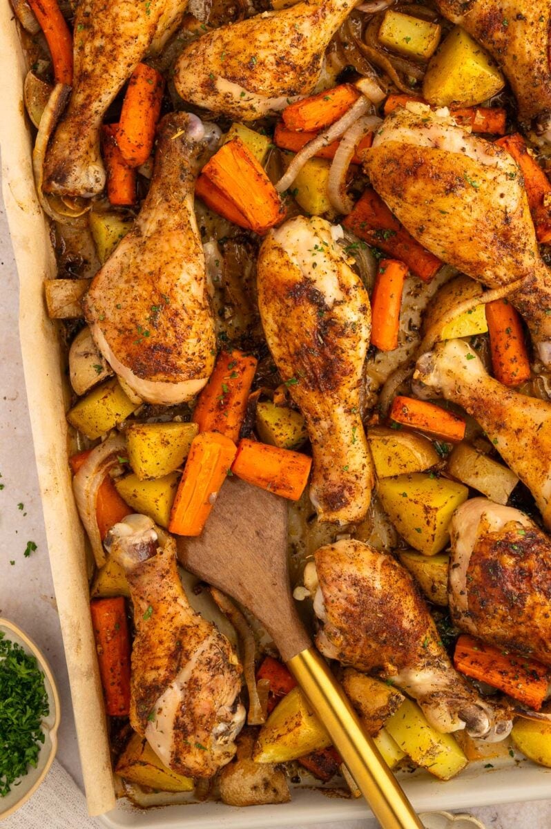 Chicken drumsticks with potatoes and carrots on a sheet pan with a spoon in them.