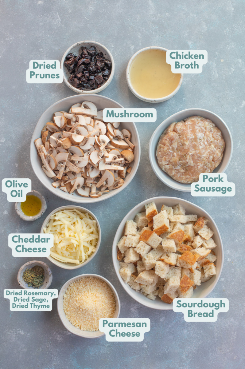 Ingredients for sourdough sausage stuffing in bowls on a grey background: chicken broth, dried prunes, mushrooms, pork sausages, olive oil, cheddar cheese, parmesan cheese, sourdough bread, dried rosemary, dried sage, dried thyme.