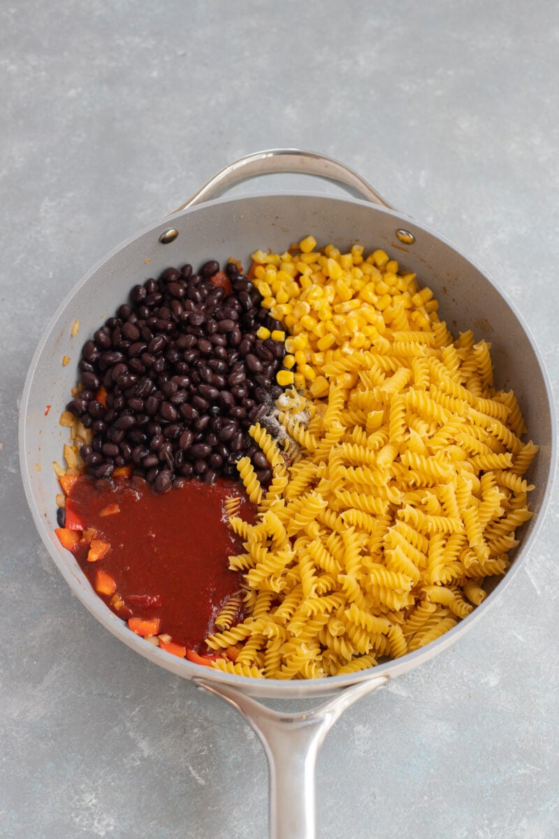 Uncooked pasta, black beans, and enchilada sauce on top of other ingredients for chicken enchilada pasta in a skillet.