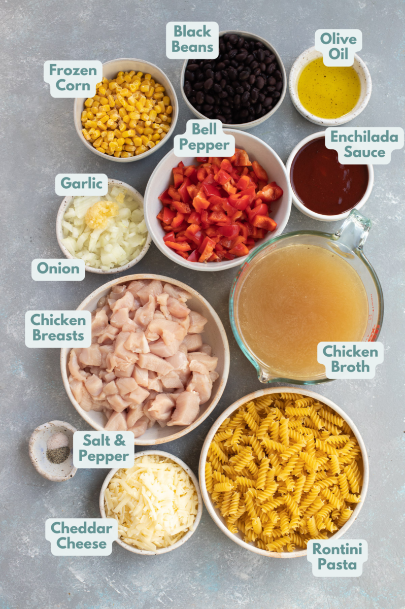 Ingredients for chicken enchilada pasta in small bowls with labels: frozen corn, black beans, olive oil, enchilada sauce, bell pepper, chicken broth, rotini pasta, cheddar cheese, salt & pepper, chicken breasts, onion, and garlic.