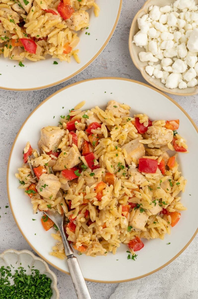 Red Pepper Chicken Orzo on a white plate with another plate and bowl of goat cheese on the side.