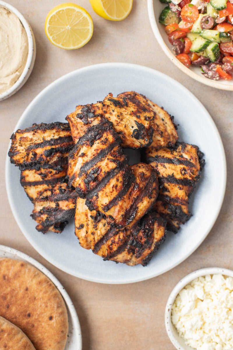 Grilled Greek Yogurt Marinated Chicken Thighs stacked on a plate with a bowl of greek salad on the side.