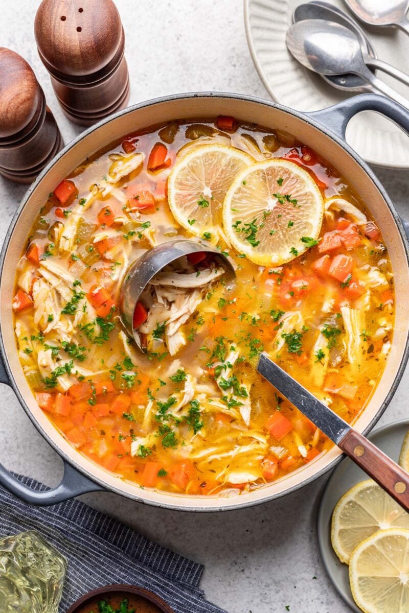 A ladle in a large pot of lemon chicken orzo soup garnished with sliced lemons.