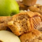 An Apple Protein Muffin with a bite taken out of it on a pile of more muffins with a green apple in the background.