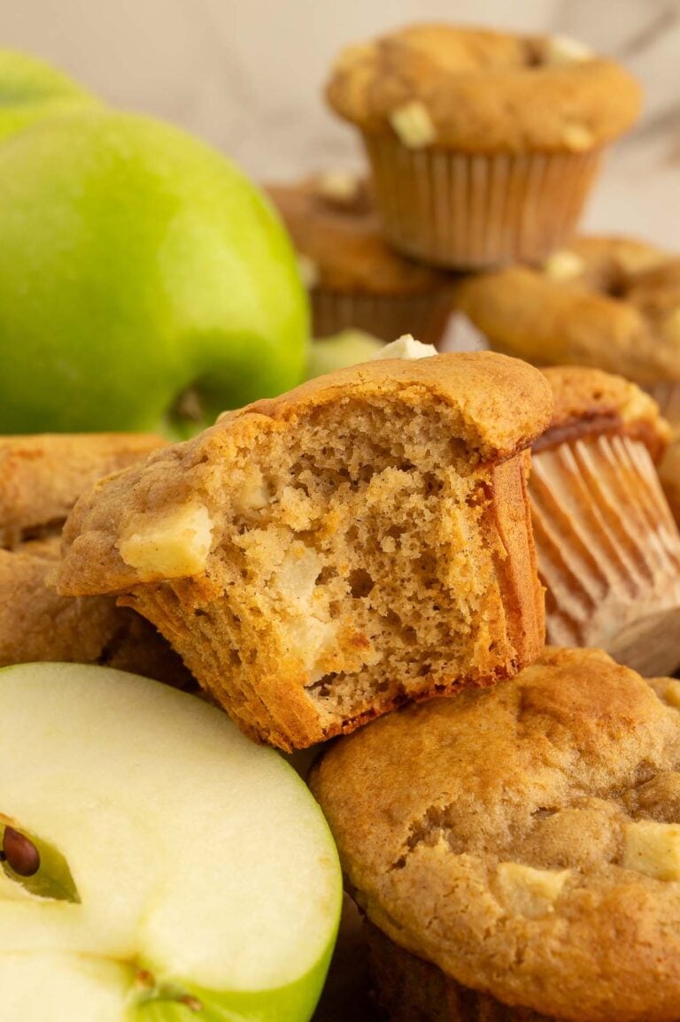 An Apple Protein Muffin with a bite taken out of it on a pile of more muffins with a green apple in the background.