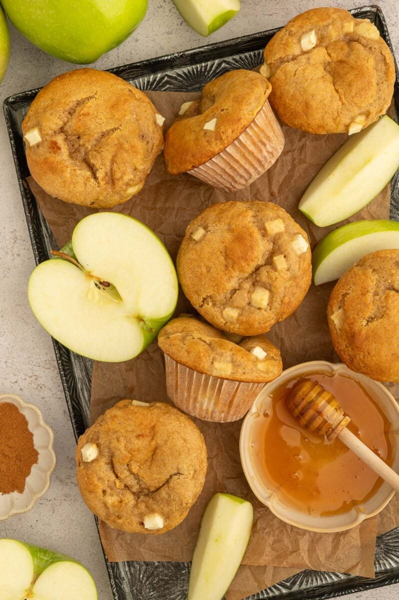 An overhead shot of apple protein muffins on a baking sheet with sliced apples scattered too.