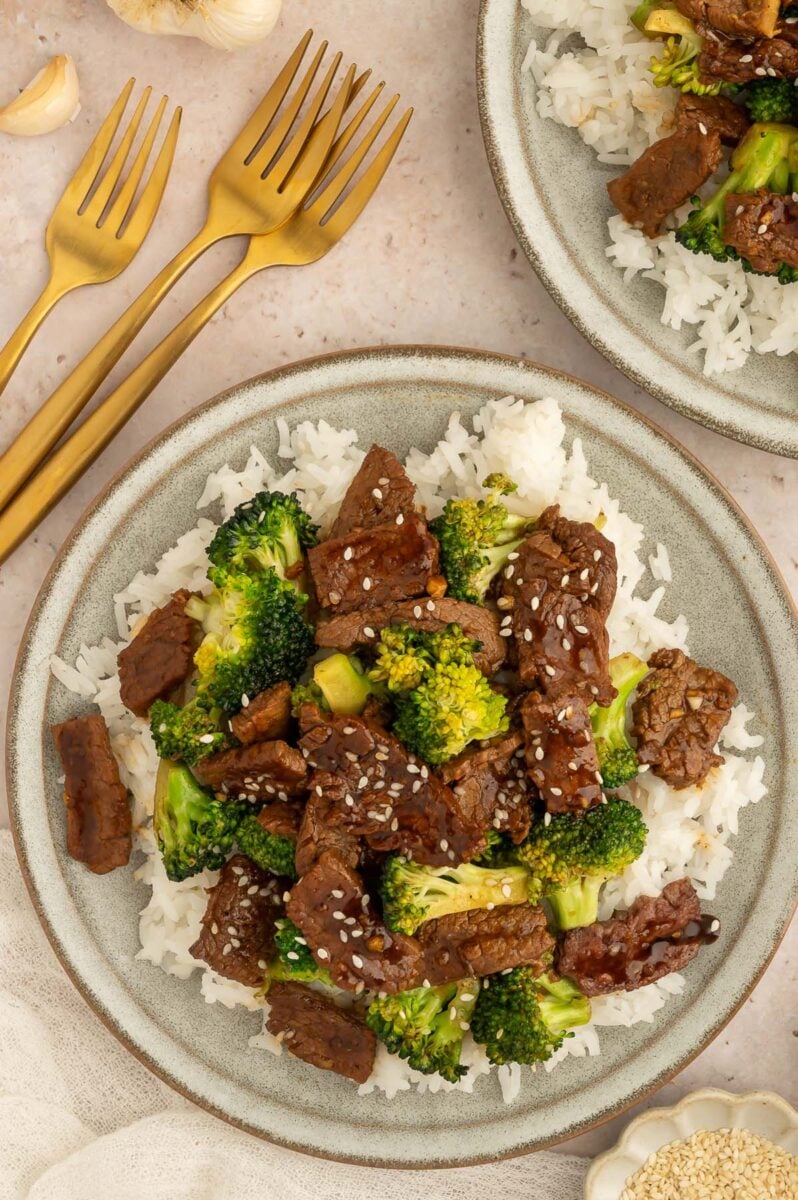 A plate of healthy beef and broccoli on a bed of white rice with forks and another plate on the side.