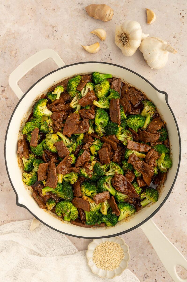 A pan of healthy beef and broccoli on a counter with garlic cloves on the side.