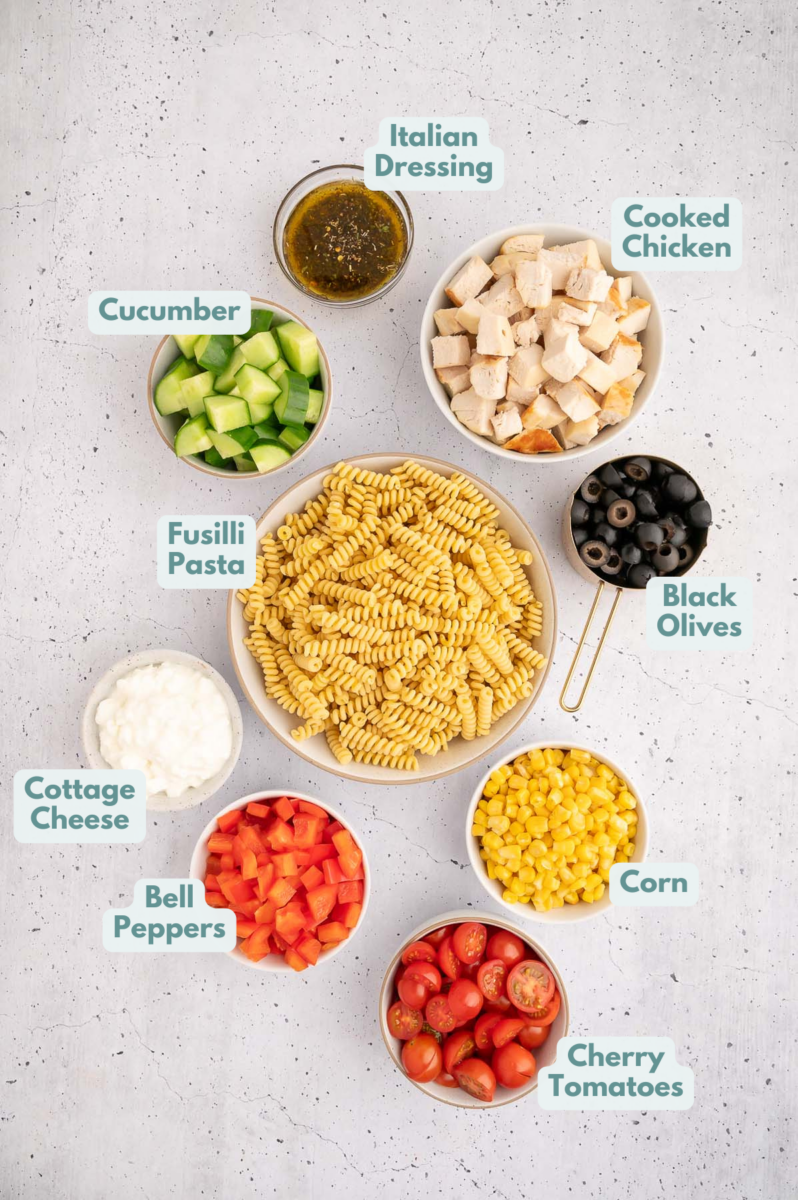 Ingredients to make high protein pasta salad in bowls and measuring cups on a counter.