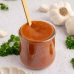 A small glass jar filled with healthy bbq sauce with a gold spoon in it with spices and garlic on the side.