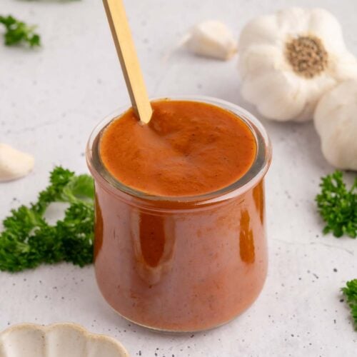A small glass jar filled with healthy bbq sauce with a gold spoon in it with spices and garlic on the side.