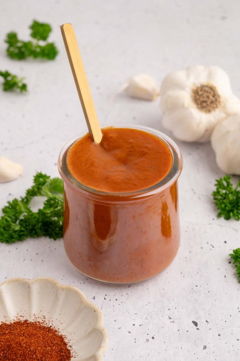 A small glass jar filled with healthy bbq sauce with a gold spoon in it with spices and garlic on the side.