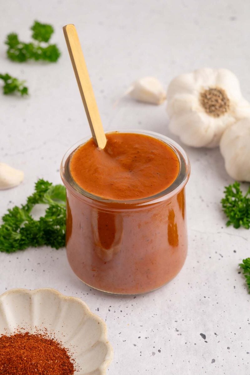 A small glass jar filled with healthy bbq sauce with a gold spoon in it with spices and garlic on the side.