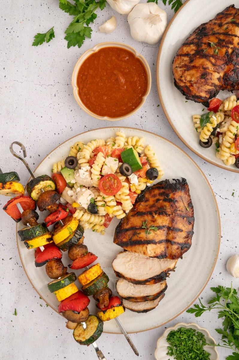 Balsamic grilled chicken on a plate with pasta salad and grilled veggie kebabs with a bowl of healthy bbq sauce on the side.