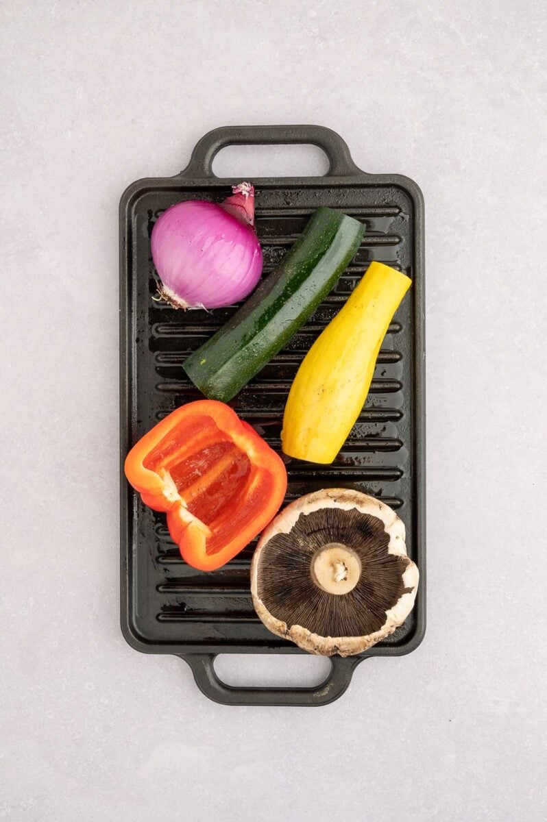 A red onion, zucchini, squash, red bell pepper, and mushroom uncooked on a grill. 