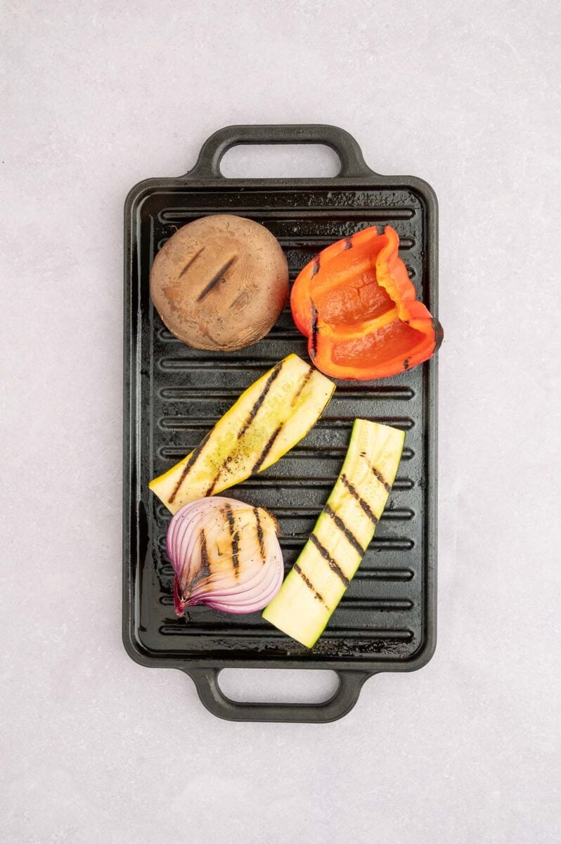 A red onion, zucchini, squash, red bell pepper, and mushroom with grill marks on a grill.