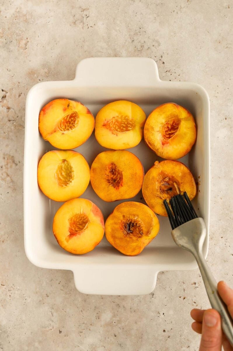 Halved peaches in a baking dish being basted with a brush.