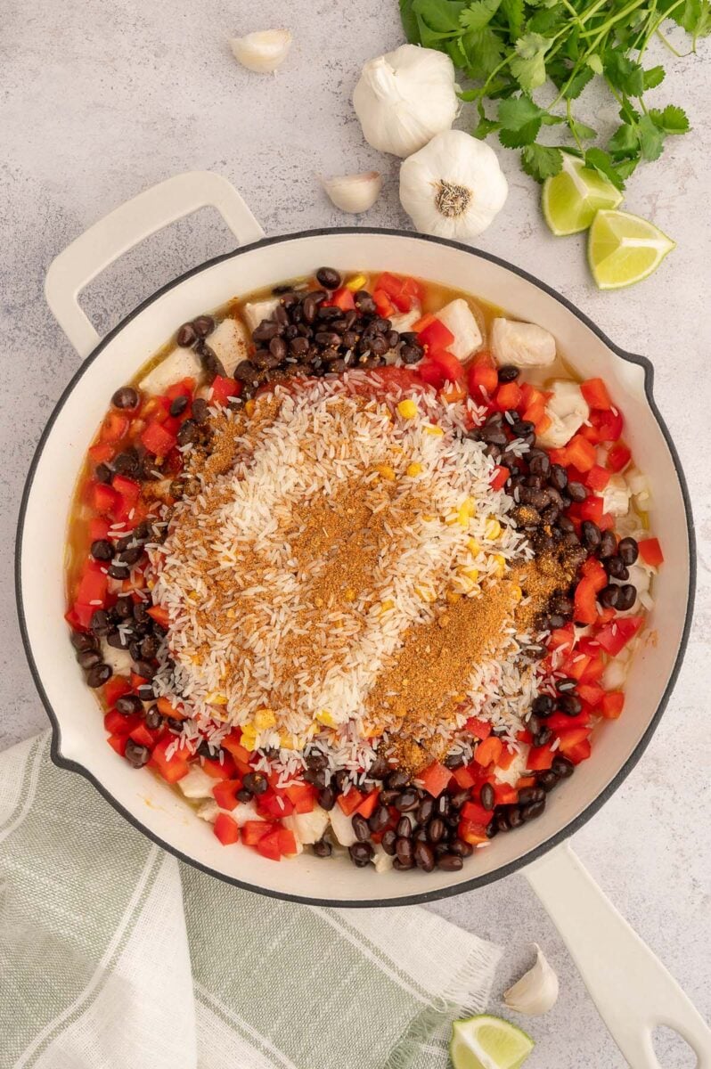Veggies, tomato sauce, chicken broth, and cubed cooked chicken in a skillet topped with uncooked rice and taco seasoning.