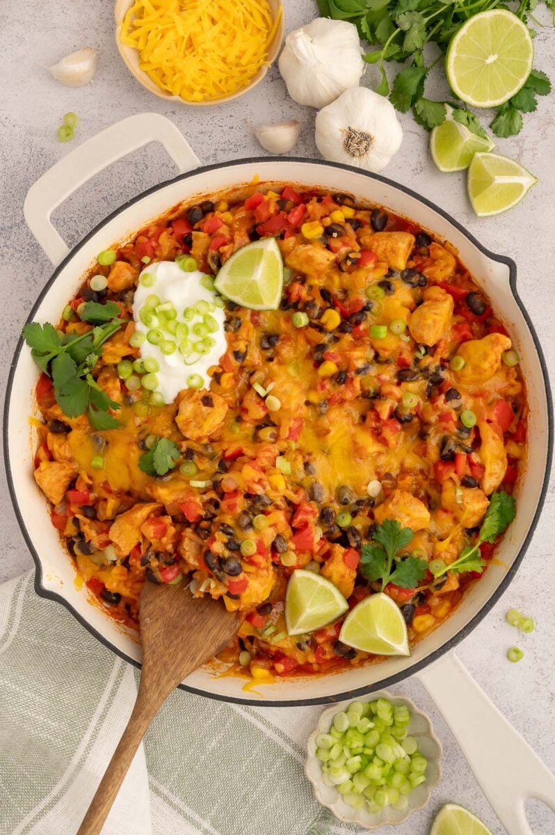 An overhead shot of cheesy chicken taco rice skillet with a wooden spoon in it with lime slices and a dollop of sour cream.