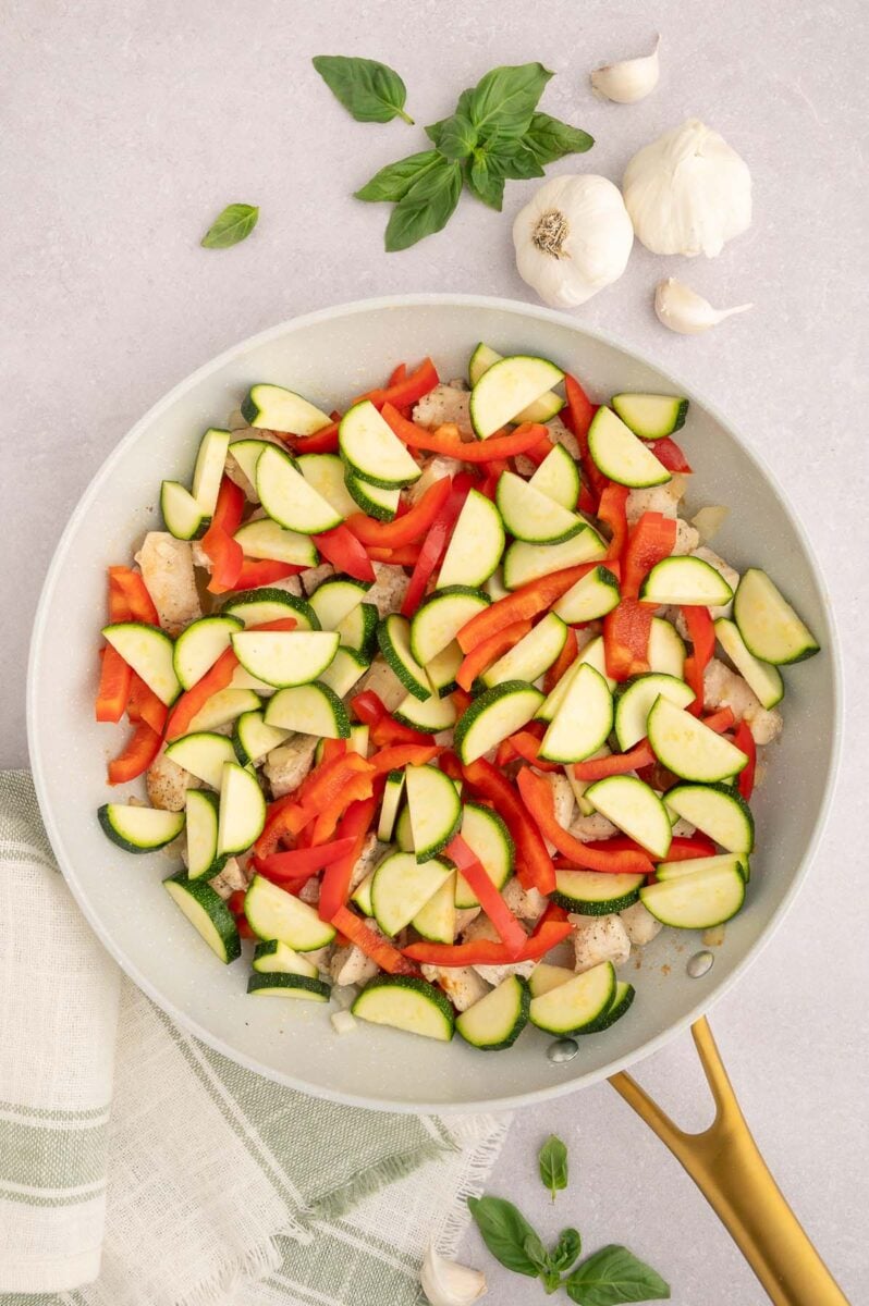 Uncooked zucchini and red bell peppers in a skillet with cooked chicken.