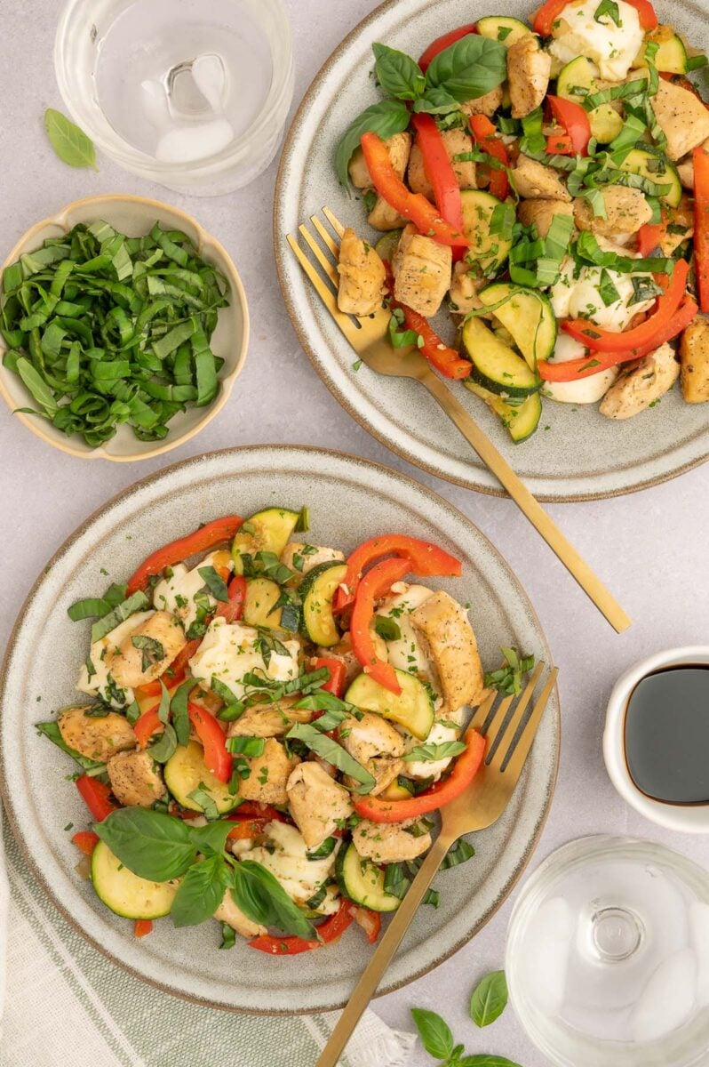 Two plates filled with chicken veggie skillet with forks on them and a bowl of chopped basil on the side.