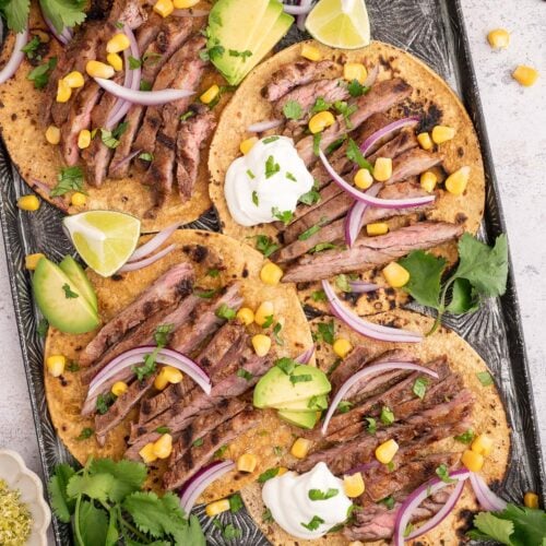 Chili Lime Steak on tacos shells garnished with red onions, cilantro, and corn on a baking sheet.