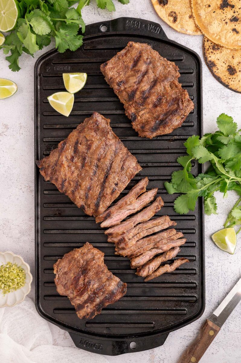Chili Lime skirt steak with grill lines on a grill pan with cilantro and limes on the side.