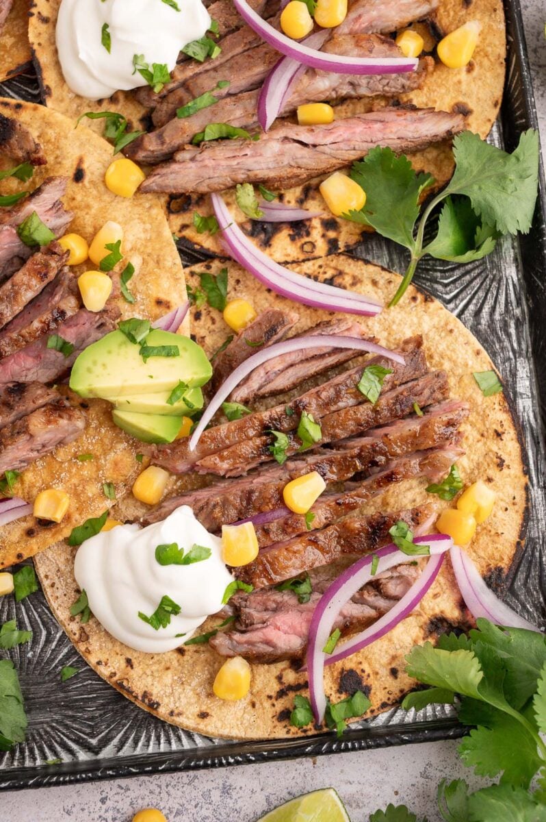 A chili lime steak taco garnished with a dollop of sour cream, corn, red onions, avocado, and cilantro.
