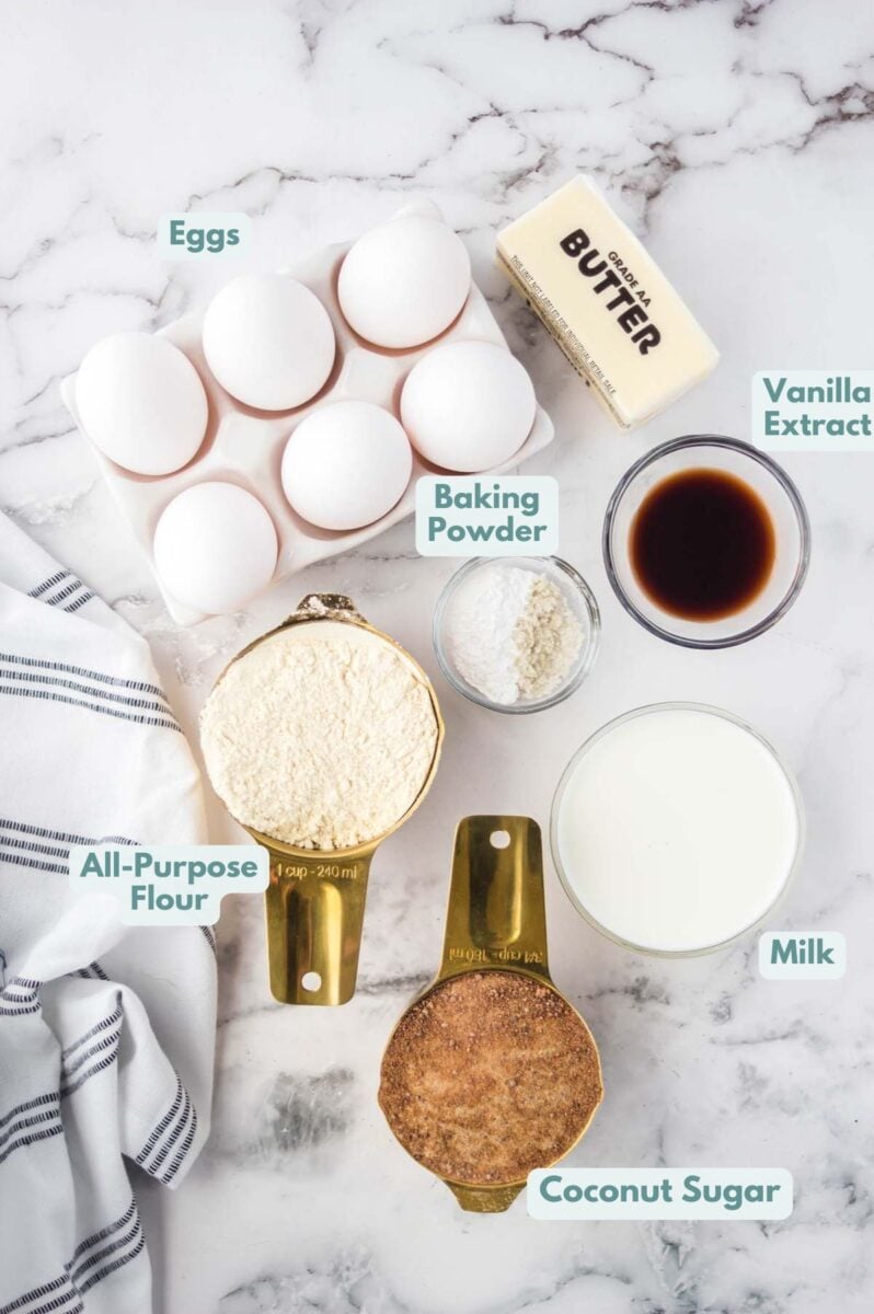 Labeled ingredients in bowls and measuring cups sitting on a marble countertop: eggs, butter, all-purpose flour, baking powder, vanilla extract, milk, and coconut sugar.