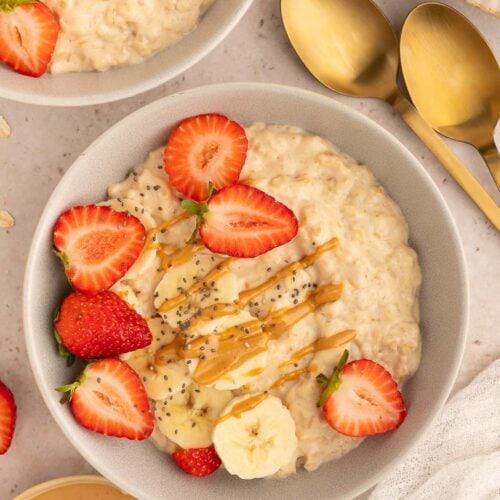 An overhead shot of a bowl of protein oatmeal with sliced strawberries and bananas and chia seeds with a drizzle of peanut butter on top and gold spoons on the side.