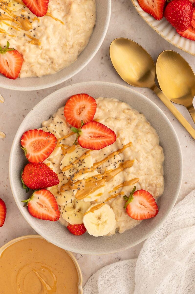 An overhead shot of a bowl of protein oatmeal with sliced strawberries and bananas and chia seeds with a drizzle of peanut butter on top and gold spoons on the side.