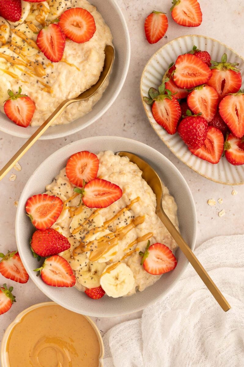 An overhead shot of two bowls of protein oatmeal with sliced strawberries and bananas and chia seeds on top with a drizzle of peanut butter.