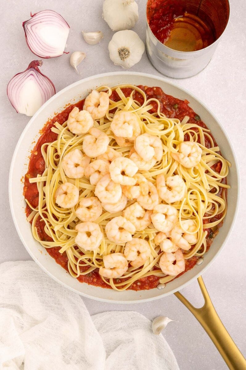 Fettuccine pasta and cooked shrimp on top of puttanesca sauce in a pan before mixing together. 