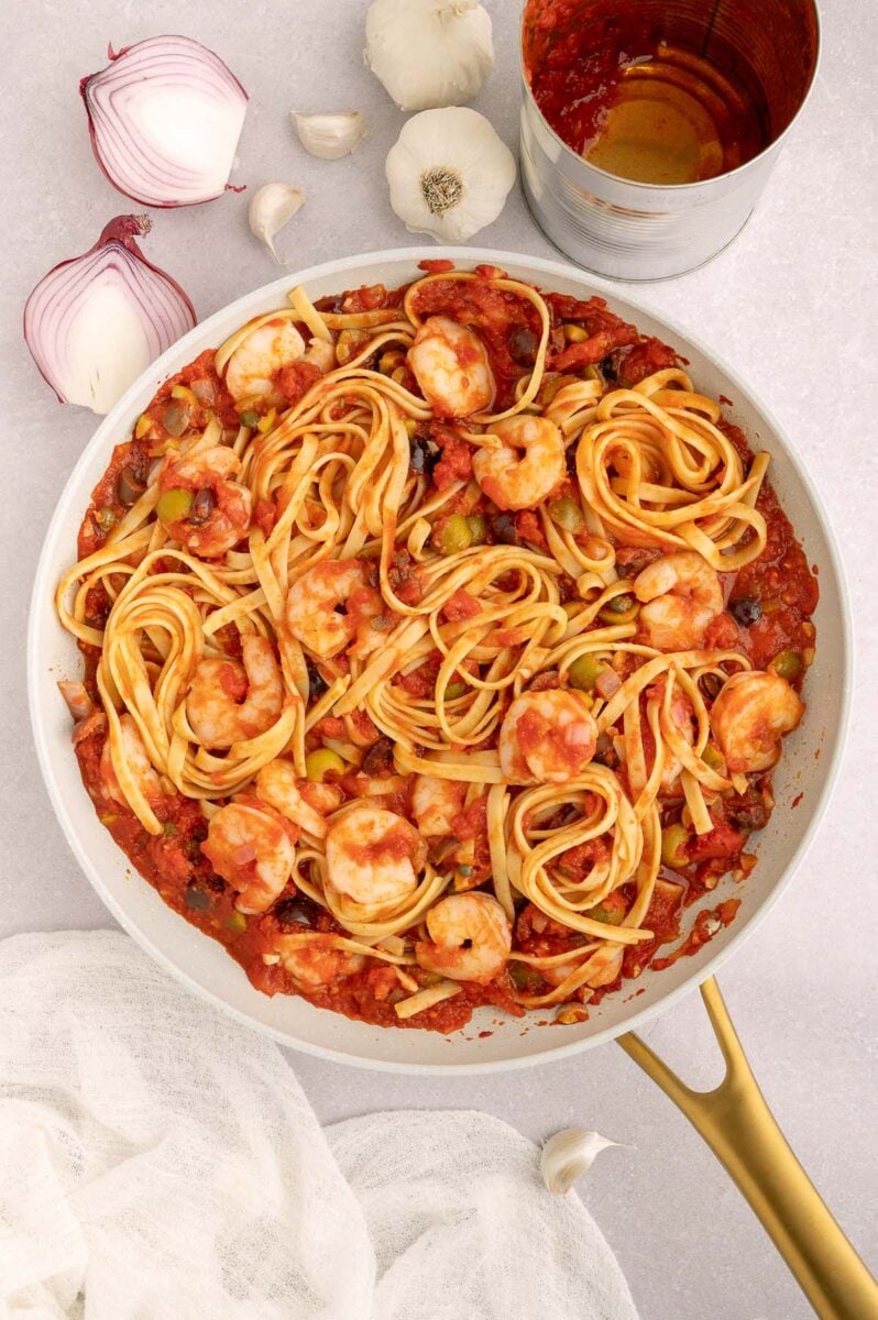 Shrimp Puttanesca in a pan on a countertop with onion, garlic and an empty tomato can on the side.