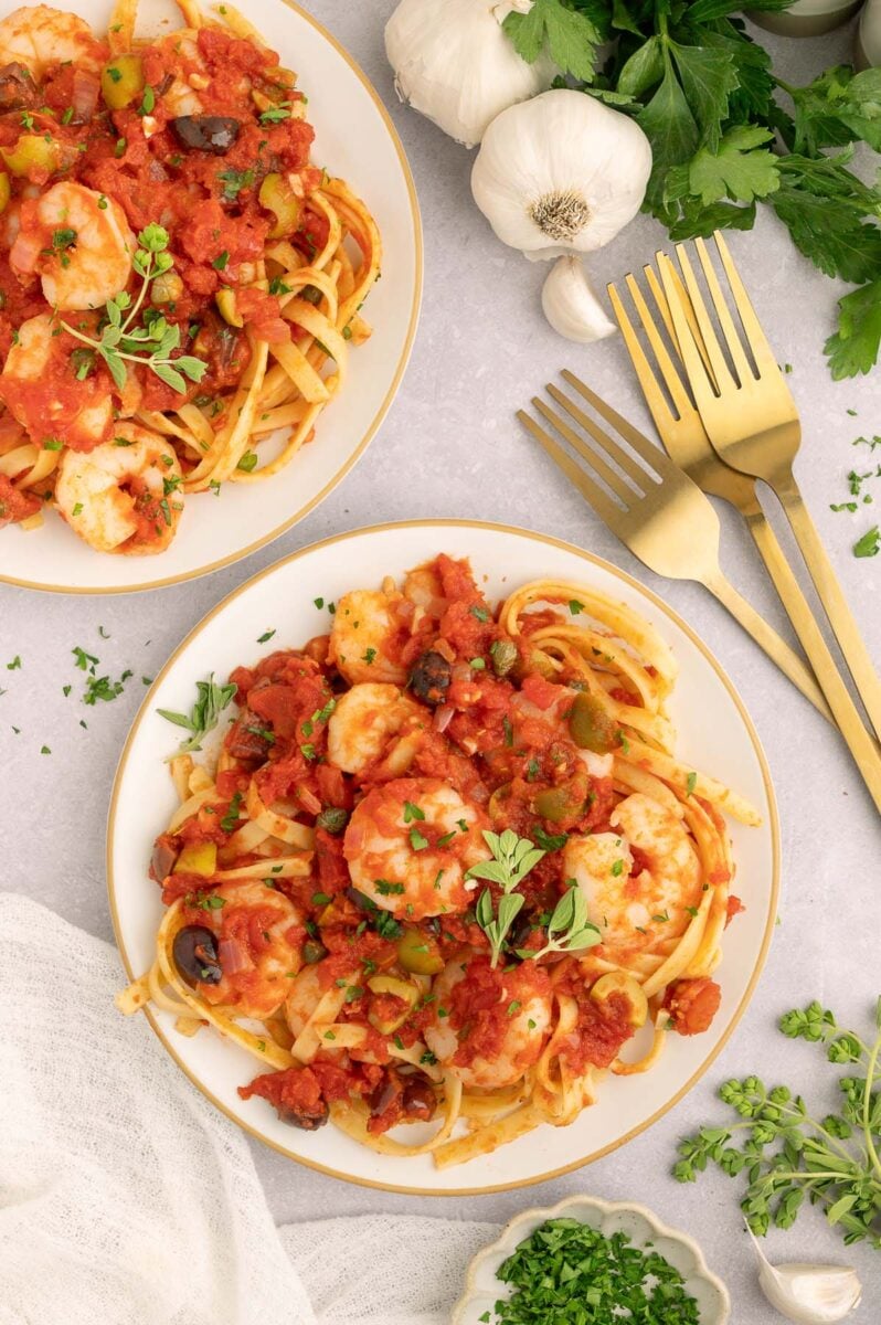 Two plates of Shrimp Puttanesca with gold forks, garlic, and herbs on the side. 