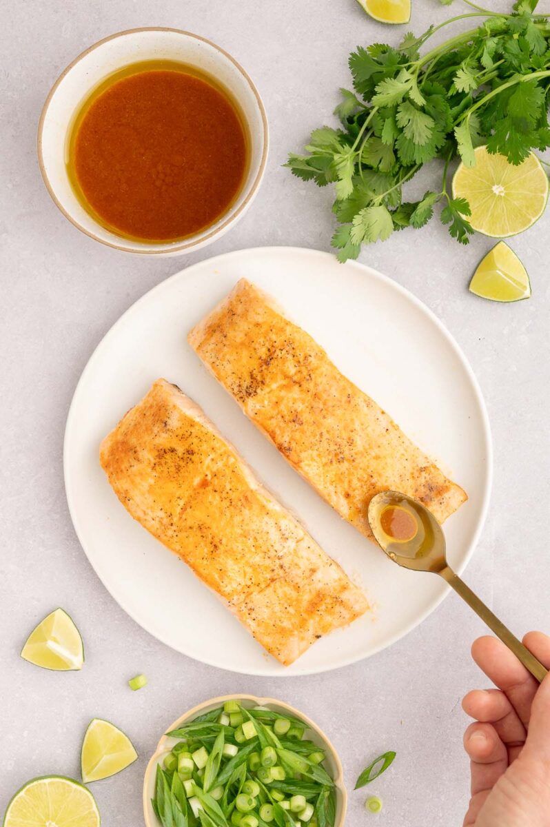 Two cooked salmon filets being drizzled with sweet chili dressing with more dressing on the side.