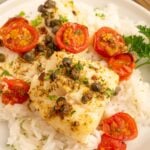 Baked cod with tomatoes and capers on a bed of white rice on a white plate.