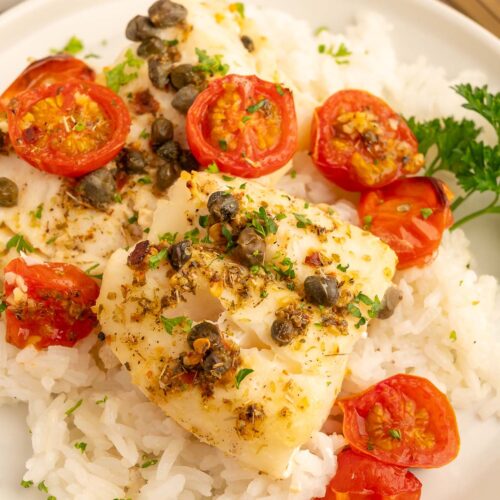 Baked cod with tomatoes and capers on a bed of white rice on a white plate.