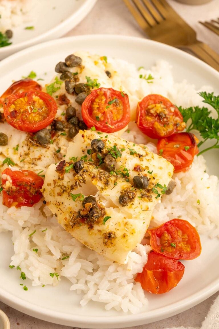 Baked cod with tomatoes and capers on a bed of white rice on a white plate.