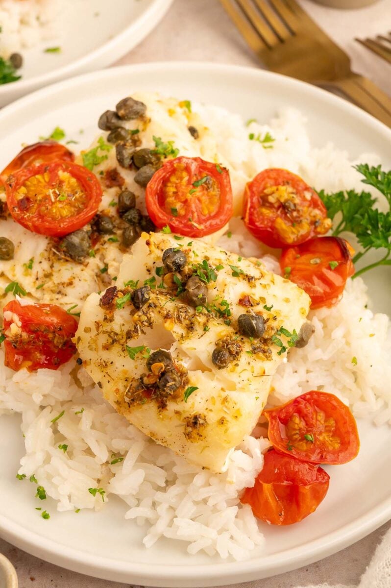Baked cod with tomatoes and capers on a bed of white rice on a white plate.