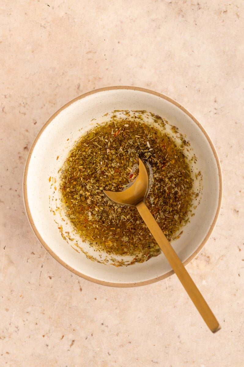 Olive oil, lemon juice, oregano, black pepper, red pepper, and salt in a small bowl mixed together with a gold spoon.