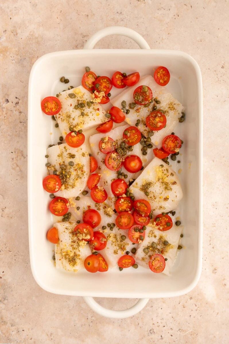 Cod filets, cherry tomatoes, capers, and minced garlic in a white baking pan before cooking.