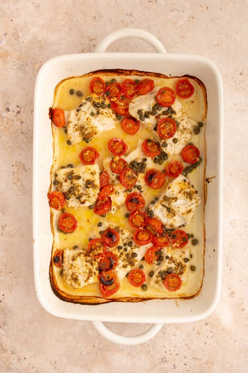 Baked cod filets with cherry tomatoes and capers in a white baking dish after cooking in the oven.