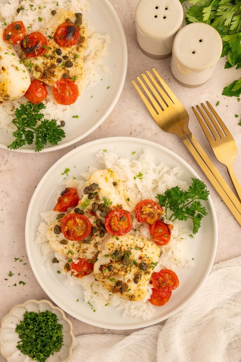 An overhead shot of two plates of baked cod with capers and cherry tomatoes on a base of white rice with gold forks on the side.