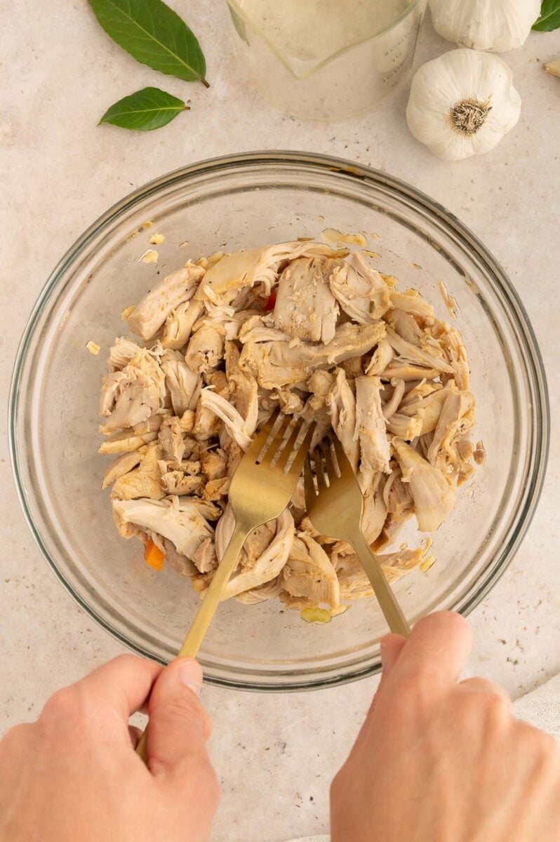 Two forks shredding chicken in a large glass bowl.